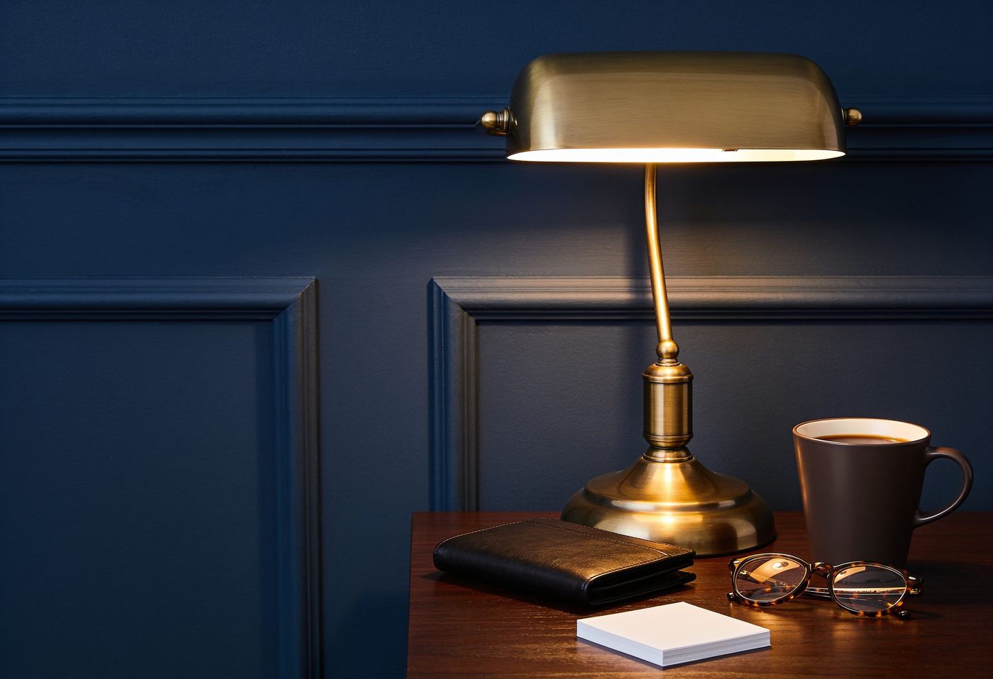 A home office desk at dusk with a brass lamp and a navy wall